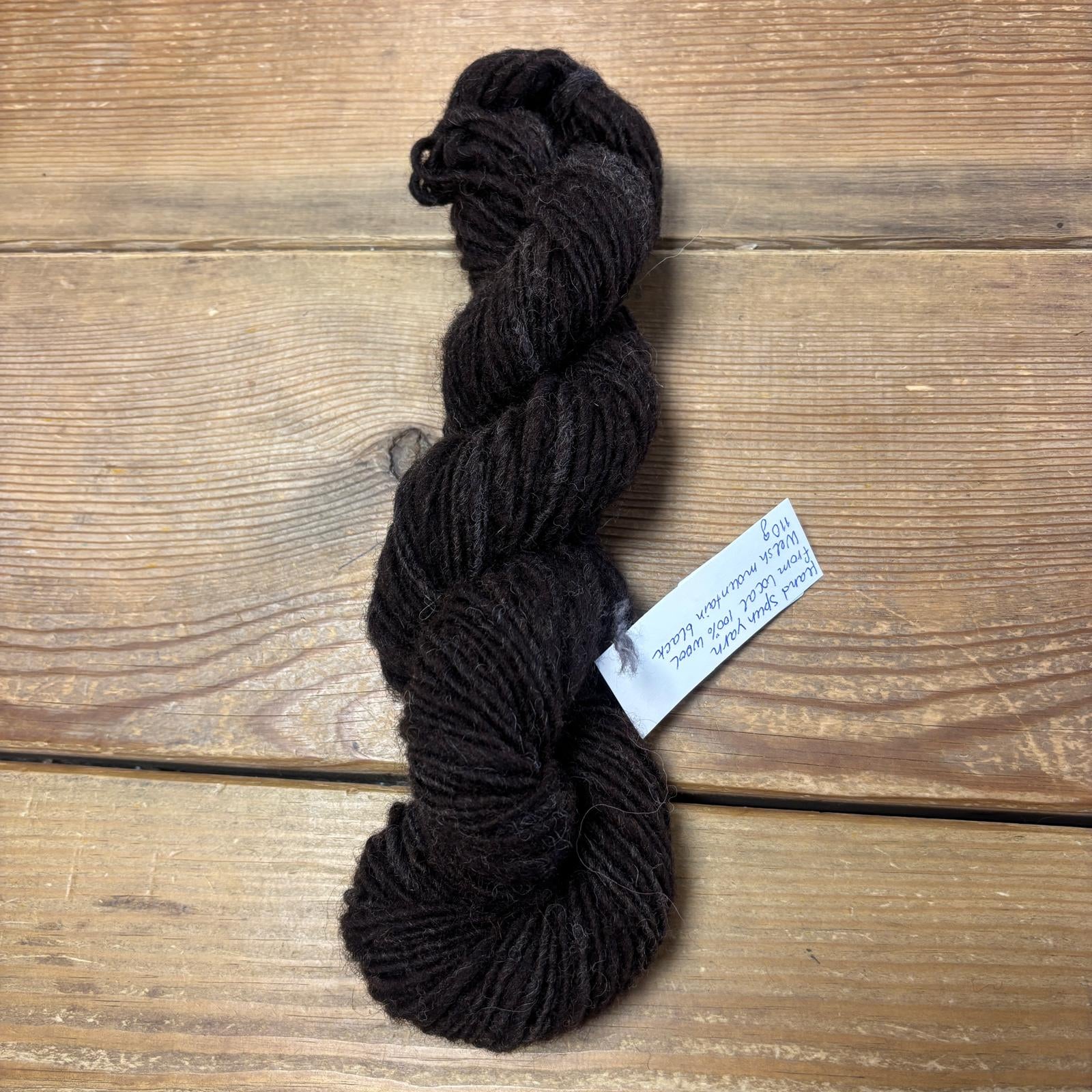 A skein of dark brown yarn with a white paper label, placed on a rustic wooden surface.